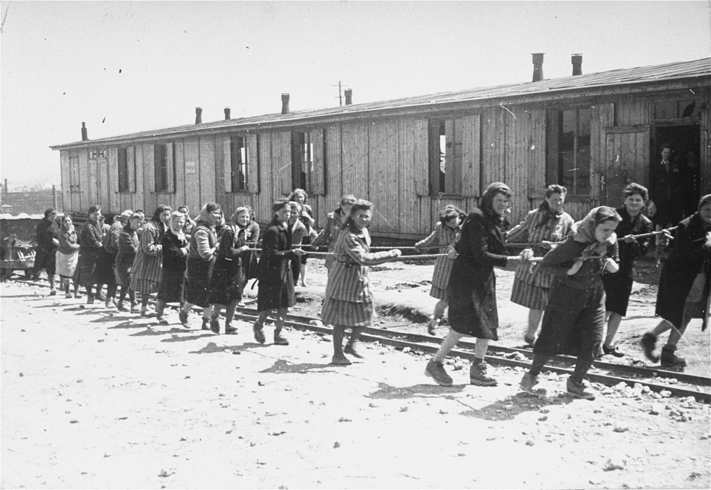 Two rows of female prisoners pull a dumpcar down a set of train tracks with two ropes. A wooden building can be seen in the background.