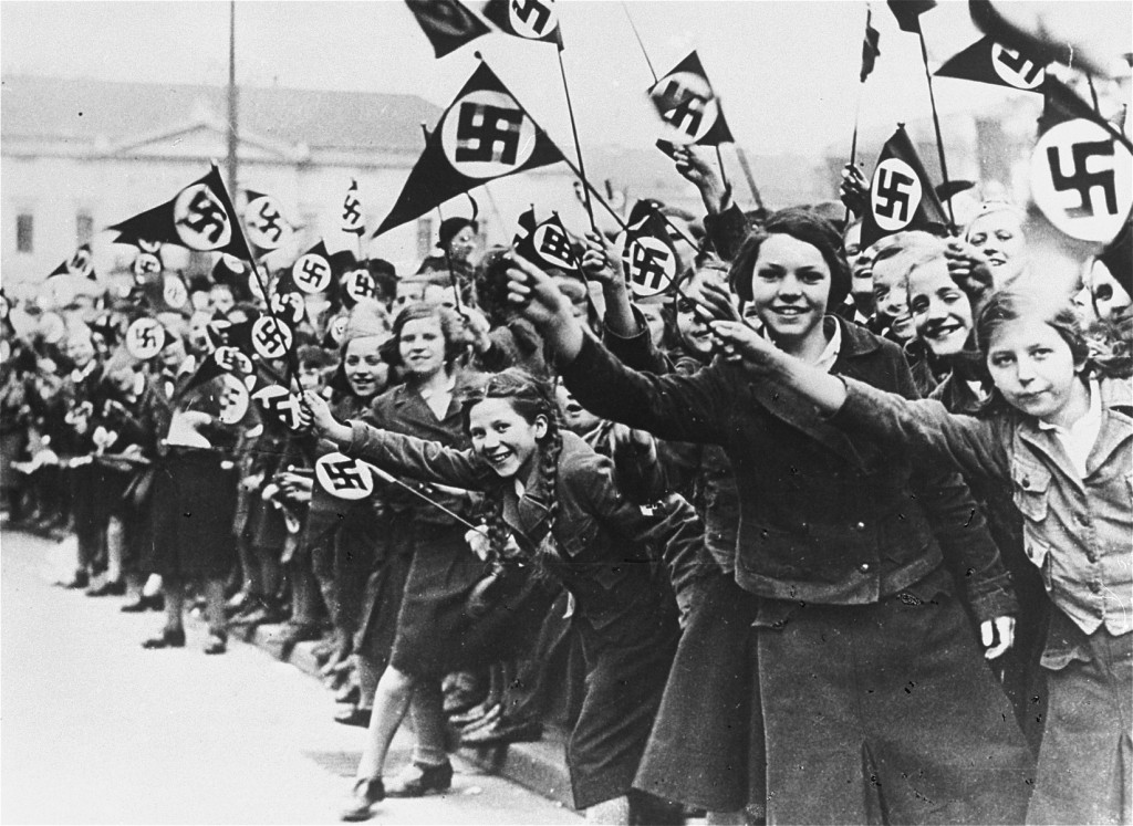 A large group of girls of various ages crowd a sidewalk waving pennant style Nazi flags.