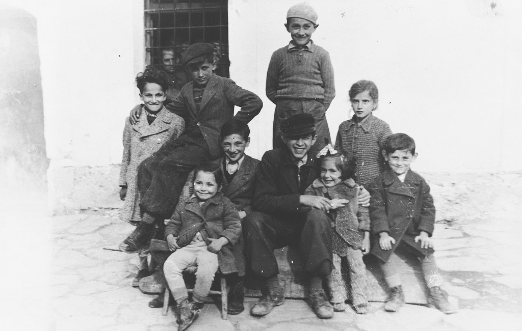 <p><span style="font-weight: 400;">Jewish refugee children interned in the prison at Pristina, circa April 1942. Gavra Mandil is seated on the right, and Irena Mandil is sitting in the front on the far left.</span></p>
<p><span style="font-weight: 400;">The Mandil family managed to escape from German-occupied Yugoslavia to the relative safety of Italian-occupied territories in present-day Kosovo. They lived freely for a short time among the Jewish community in the city of Pristina. However, Italian occupation forces soon interned the growing number of Jewish refugees in an empty school before relocating them to the city’s prison. The Mandils were confined there with more than a hundred others for several months. Gavra Mandil would later recall that he and his sister Irena spent much of their time at Pristina playing war in the prison courtyard with the other children.</span></p>
<p><span style="font-weight: 400;">The Italian guards treated the interned Jews decently. Mosa Mandil began to earn their respect and favor with his photography skills. He created portraits of the guards and officers and soon was on friendly terms with them. But German authorities were pressuring the Italians to turn over Jewish refugees to them. German forces took custody of dozens of the interned Jews at Pristina in late spring 1942 and killed them. In response, the Italian guards at Pristina prison found several trucks to transport the remaining Jews farther away from German influence. The Jewish refugees drove to Italian-occupied Albania. Each truck went to a different city or town. The Mandils and several other families went to the Albanian city of Kavaja. </span></p>
