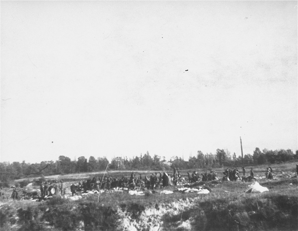A large crowd of people stand in the middle of a field in various states of undress. Piles of possessions are scattered across the field. A few unformed men can be seen threatening the crowd.