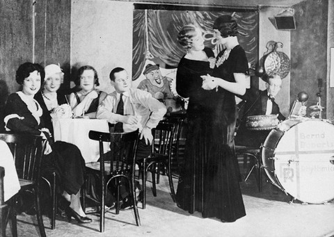 Two women with light skin tone in dark dresses and styled hair dance together inside of a nightclub. Part of a drum kit sits to the right of the dance floor while several people sit in tables to the left.