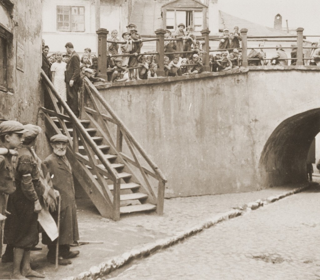 Several people stand along an elevated walkway leading towards a bridge. A wooden staircase has been added to the walkway, providing access to the road below. A few individuals stand along the side of the lower road.