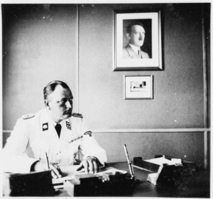 The commander of Gross-Rosen, SS-Obersturmbannfuehrer Arthur Roedl, at his desk with a photograph of Adolf Hitler hanging on the wall.
