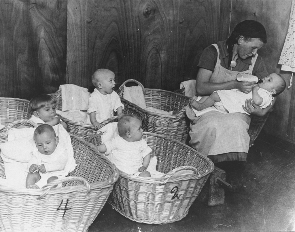 A woman with medium-light skin tone and long hair sits bottle feeding a baby with five woven baskets next to her. Four other babies of various ages sit in the open numbered baskets. All of the babies have light skin tones.