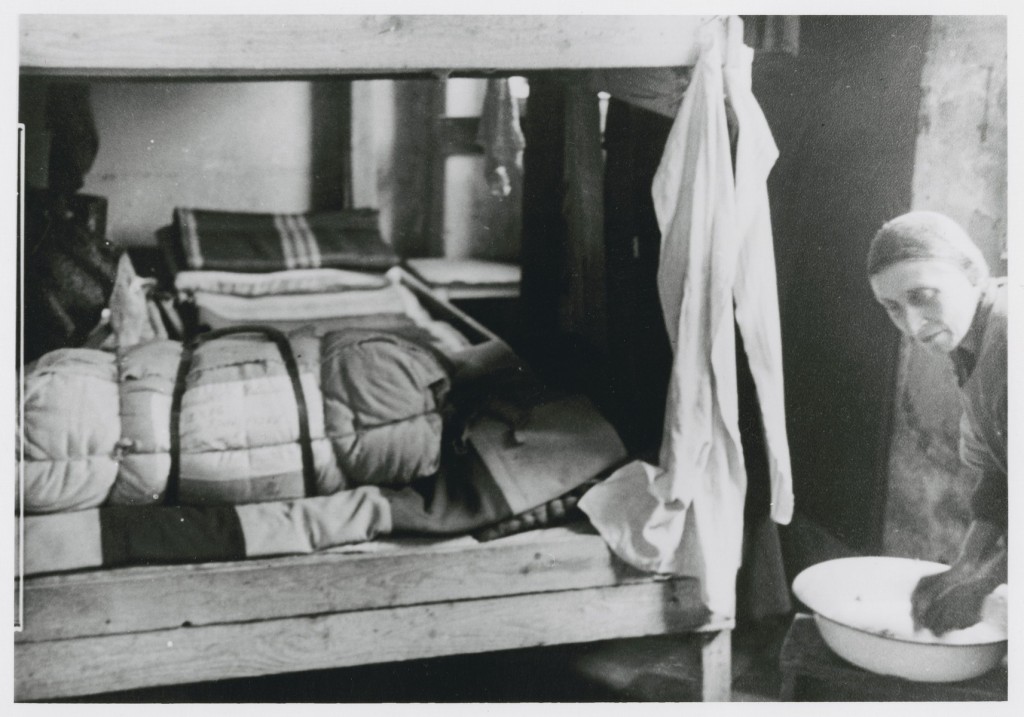 Two wooden bunks fill a small room. An older woman with light skin tone and wearing a head covering stands to the right, washing a rag out in a basin.