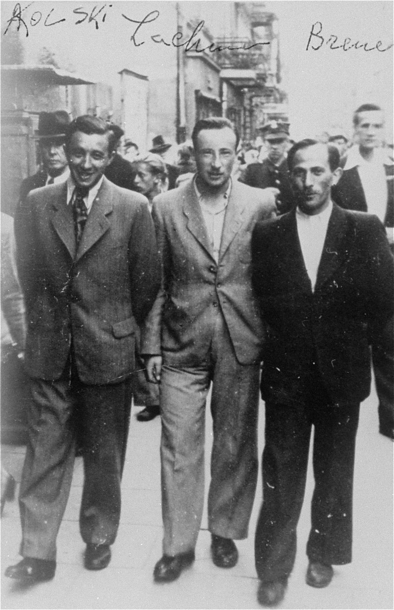 Three men walk together down the middle of a busy street. Each one is wearing a suit of a different color.