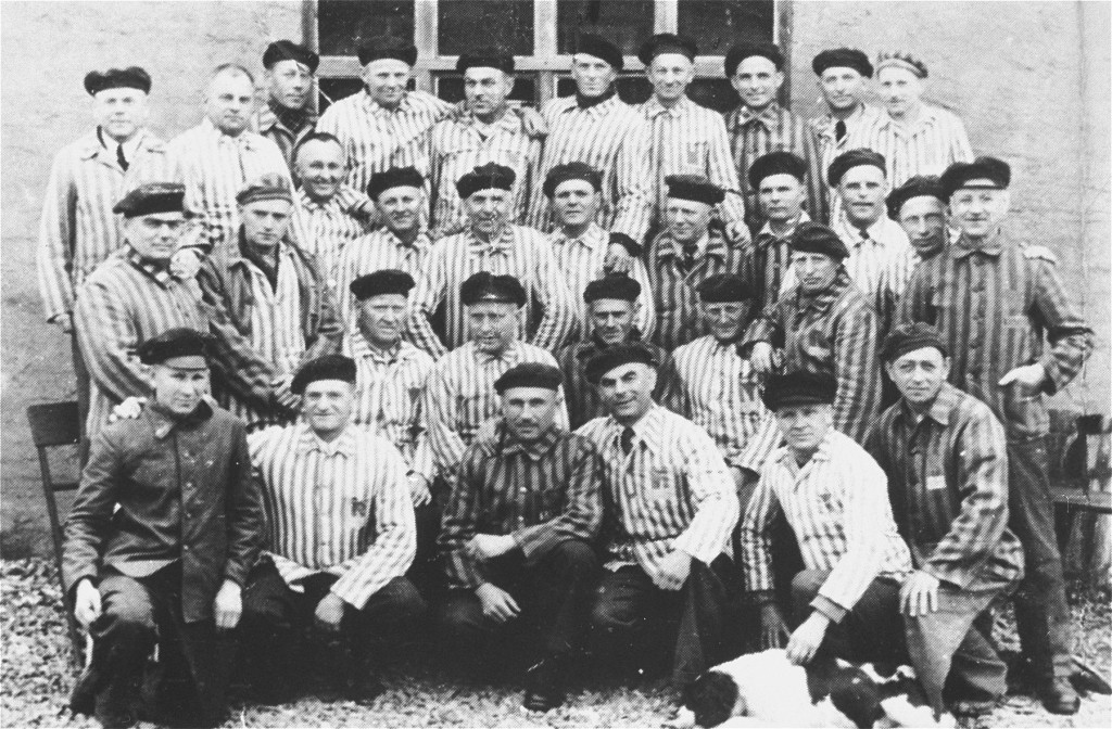 A large group of men in rows pose for a photograph. Most of the men are wearing striped uniforms and dark caps. There are 32 people total.