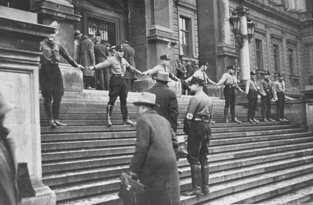 A group of uniformed Nazis stand in a line with their arms spread creating a barrier on the steps of a large building blocking individuals from entering the building. A couple of people stand on the steps below the human barricade, looking up towards them.