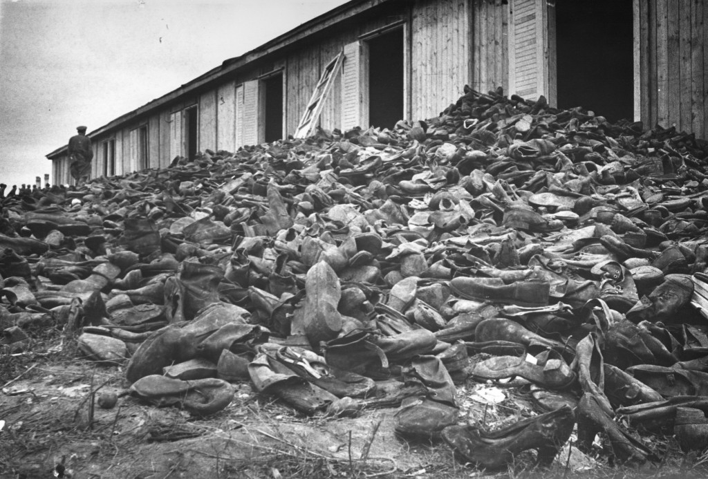A uniformed soldier stands atop a mound of shoes outside of a large warehouse. Hundreds of shoes of every size and shape come up to the middle of the warehouse doors and appear to go on in perpetuity.