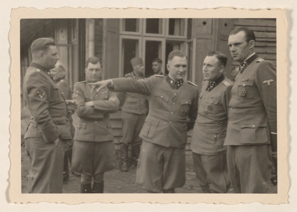 Four S-S officers stand around with other uniformed soldiers. The second from the left is pointing towards the left while talking to the officer closest to him.