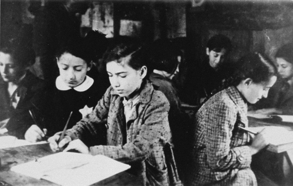 Black and white photograph of children writing with books beside them. In the background, a teacher stands and watches the children work. One of the children can be seen wearing the compulsory Star of David badge on their clothing.