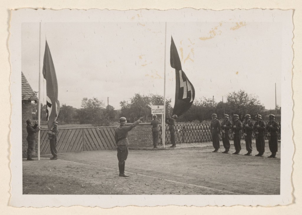 A Nazi soldier salutes as the Nazi and SS flags are raised while a line of troops stand with rifles at attention during the dedication.