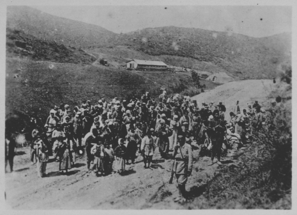 A large group of people walk down a country road with a few visible uniformed guards along the edges of the group. The photograph has been taken from a taller vantage point and the countryside is visible behind the group.