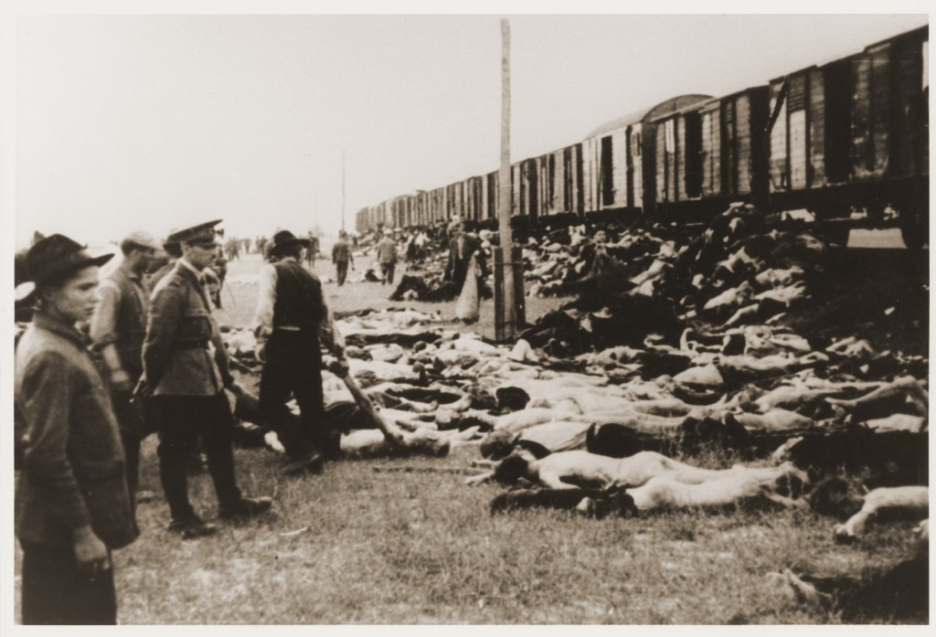A small group of people look up towards a train of open railcars. Bodies of victims stack up outside the railcars and on the grass.