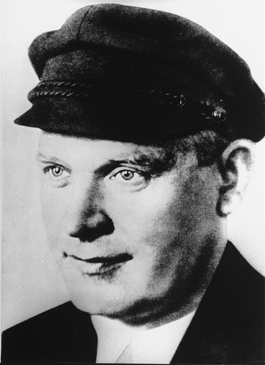 A black and white portrait of an adult man with light skin tone and short dark hair. He looks towards the left while wearing a dark cap and jacket.