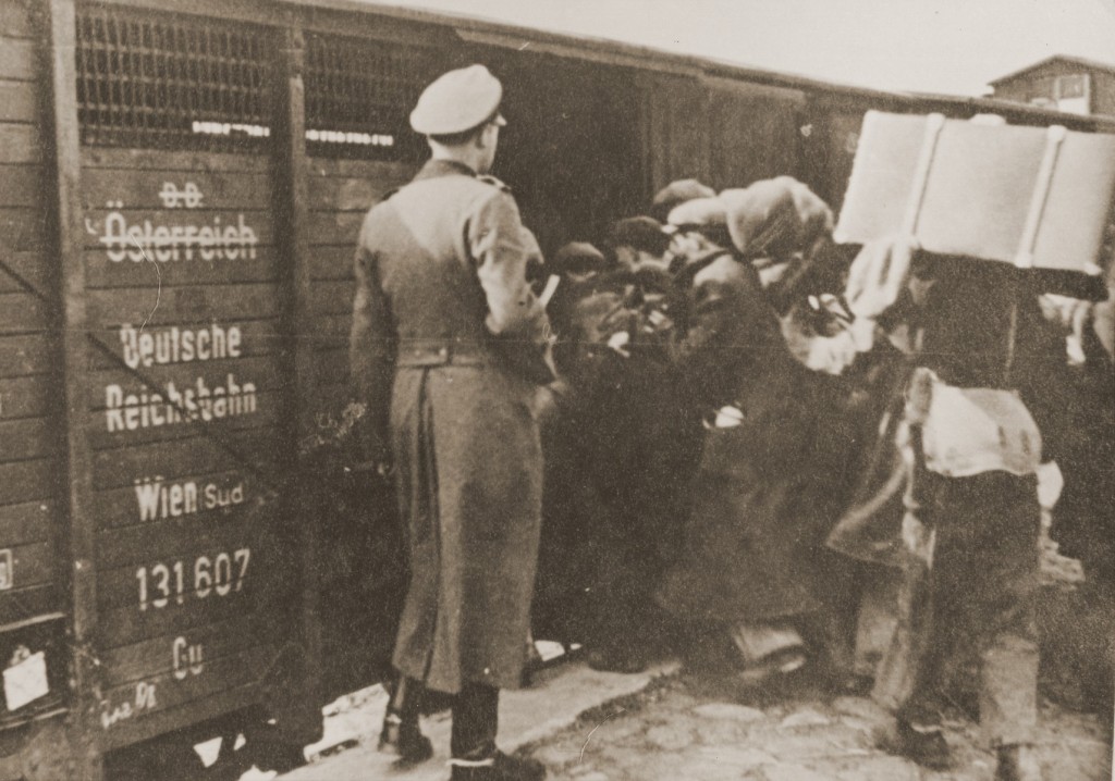 A line of people walk into a boxcar, holding bags, suitcases, and other personal belongings. A uniformed soldier stands to the left of the boxcar door supervising with his back facing the camera. The boxcar has German text on the side.