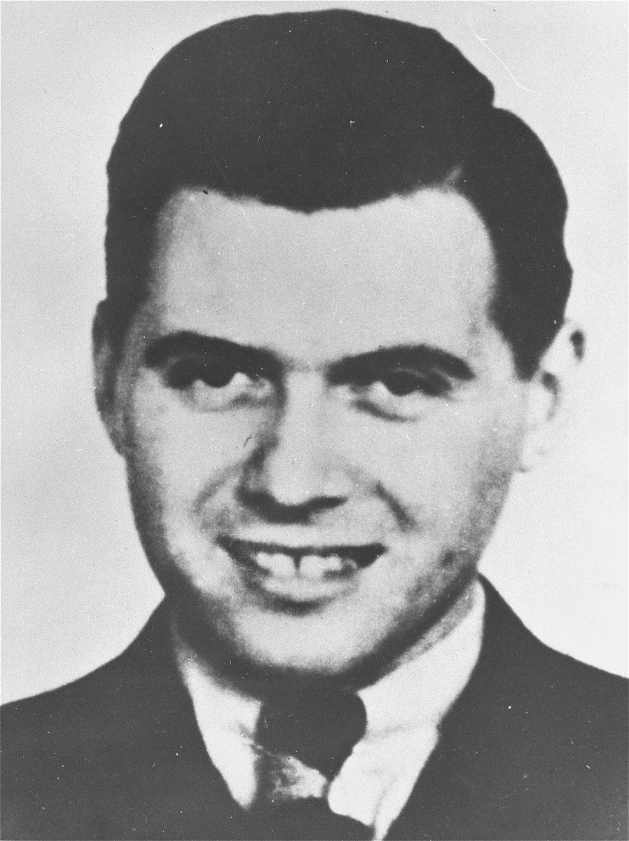 Josef Mengele grins at the camera. He is wearing a dark suit, white shirt, and tie.