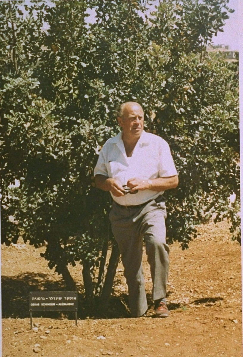Oskar Schindler stands in front of a small but mature tree looking off to the side with his hands clasped in front of him. He is wearing a white shirt with dark pants.
