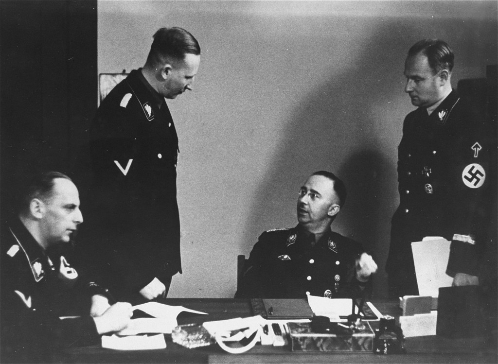 Heinrich Himmler sits at a desk while Reinhard Heydrich and another man with short dark hair and light skin tone stand on either side. Another man with short slicked back hair and light skin tone sits at the left edge of the table facing right. All are in dark military uniforms.