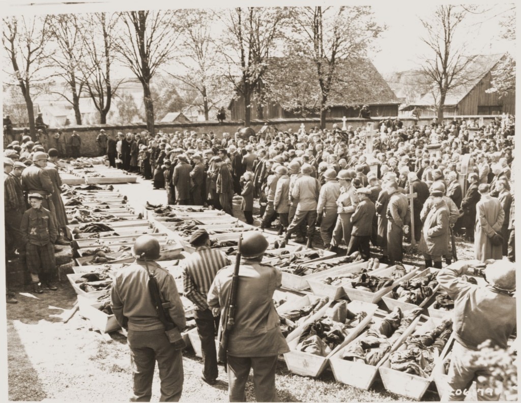 Several dozen open caskets have been lined up on the ground. Lines of civilians and soldiers walk past the caskets, while others in the crowd observe behind the procession.