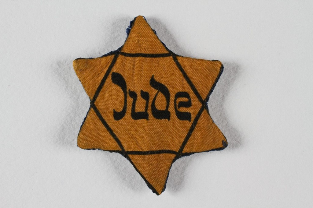 A cloth, yellow six-pointed Star of David badge with a black outline. In the center of the star is the word ‘Jude’ in black letters.