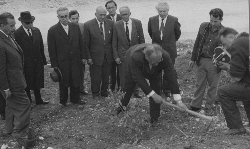 An older man in a suit uses a farm tool to plant a tree. A group of men surround him, including one man filming the entire scene.