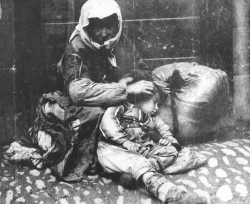 An Armenian woman and her child sit on a sidewalk next to a bundle of their possessions.