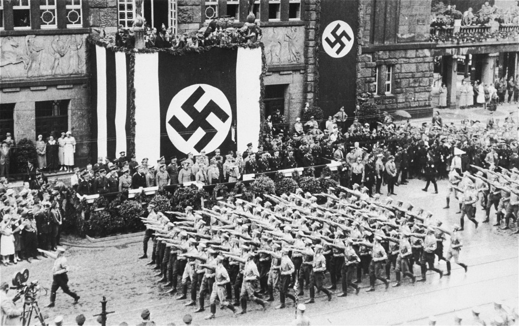 Photograph of a parade from a high view point, possibly a balcony. Adolf Hitler stands on a balcony decorated with a large swastika flag. Battalions of uniformed individuals salute Hitler as they pass by. There is a crowd of people watching, in addition to a film crew in the bottom left corner.