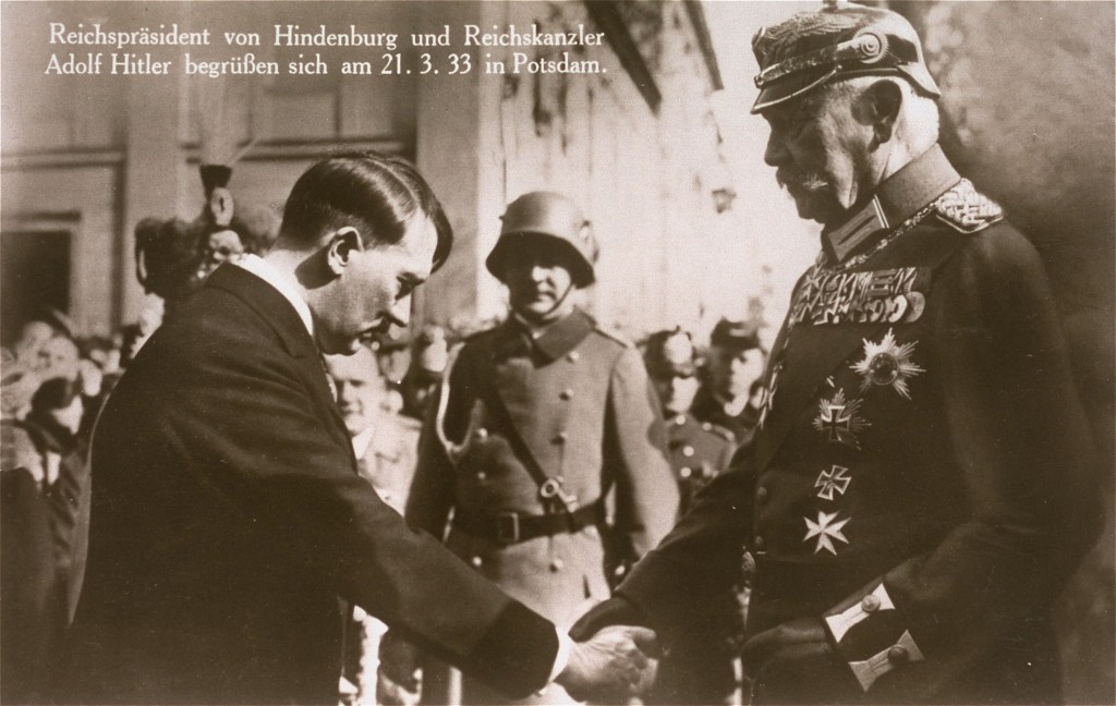 Adolf Hitler and Paul von Hindenburg greet each other surrounded by a crowd of people. The sepia toned postcard has white German text in the top left corner that reads ‘Reichspräsident von Hindenburg und Reichskanzler Adolf Hitler begrüßen sich am 21.3.33 in Potsdam.’