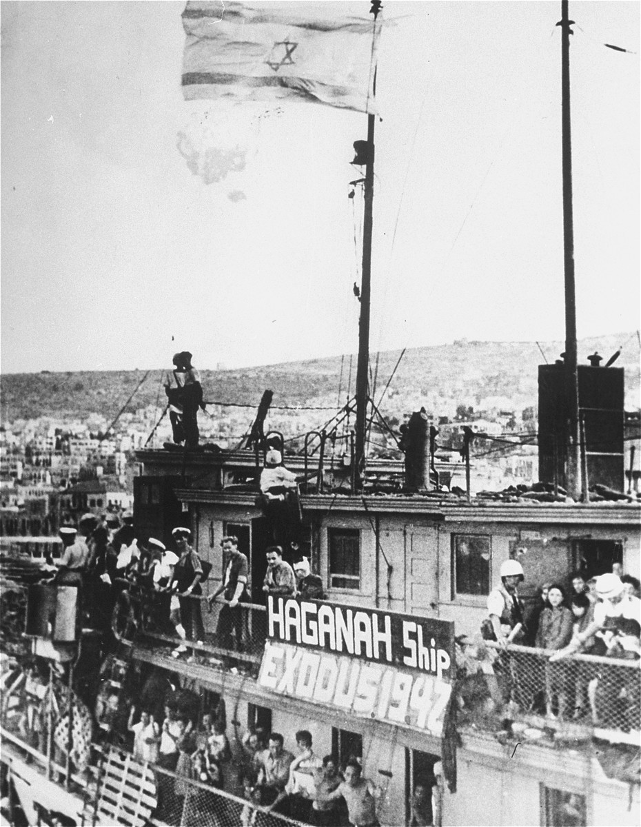 Jewish refugees and uniformed police gather along the railings of a ship. There is a large sign along the railing that reads, “Haganah Ship. Exodus 1942.”