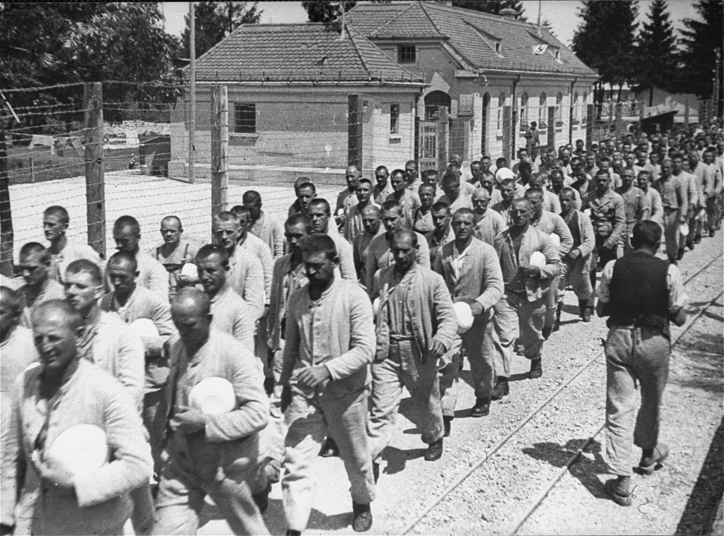 Long lines of prisoners walk next to a barbed wire fence and a large building. Many of the prisoners are in plain uniforms and are carrying light colored bowls. One person, possibly a guard, walks in the opposite direction.