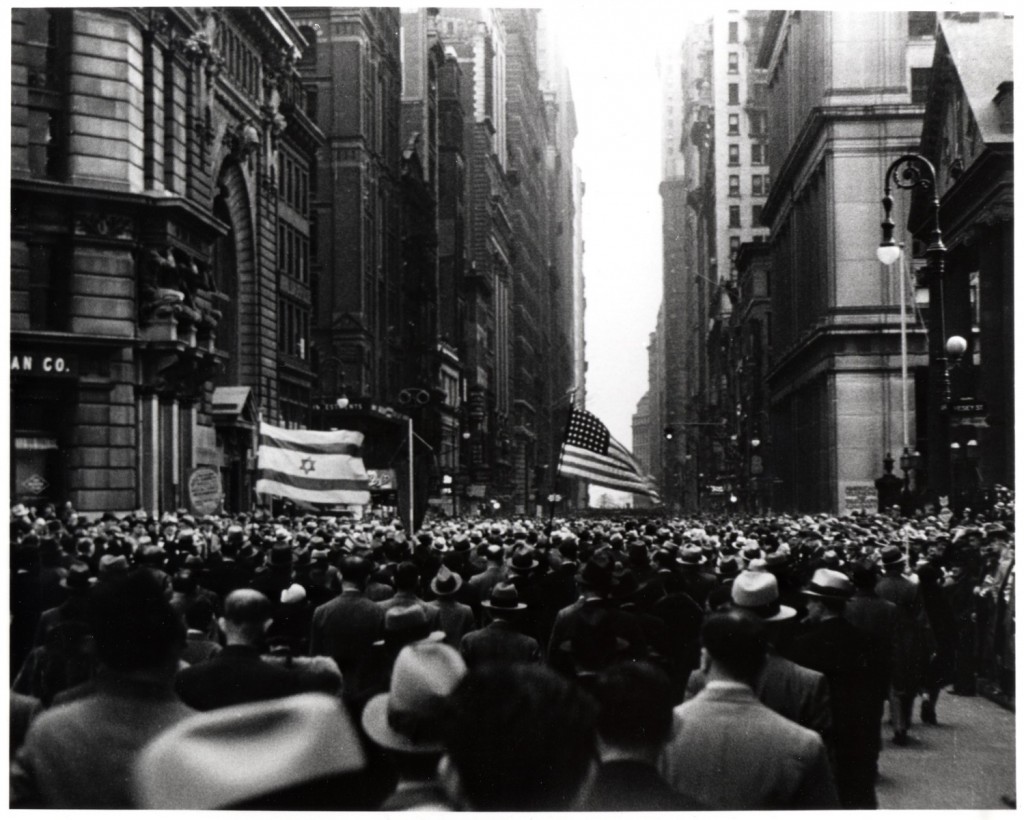 A large crowd of people gather to protest. The crowd carries two flags: the flag of Israel and the American flag.