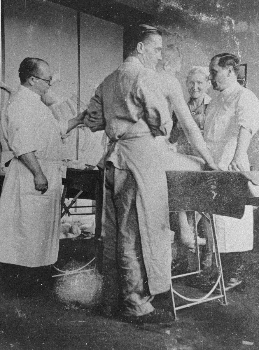 A group of medical personnel stand around a table examining a nude patient. Two men stand on one side of the table closest to the camera, while one man and one woman stand on the other.
