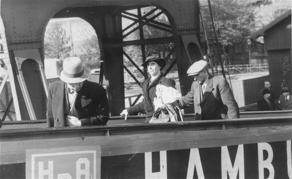 Three individuals with light skin tone walk up a walkway to board a ship. A large metal structure can be seen in the background.