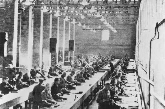 The inside of a large factory with tables in rows. Rows of prisoners sit on one side of the tables and work on materials in front of them.