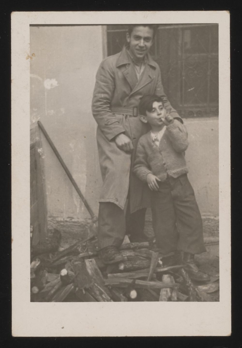 Wearing warmer clothing, a young man and young boy with light skin tones and dark hair stand on a pile of chopped wood. The boy has his thumb in his mouth and leans against the man. The man smiles and rests his hand on the boy’s shoulder.