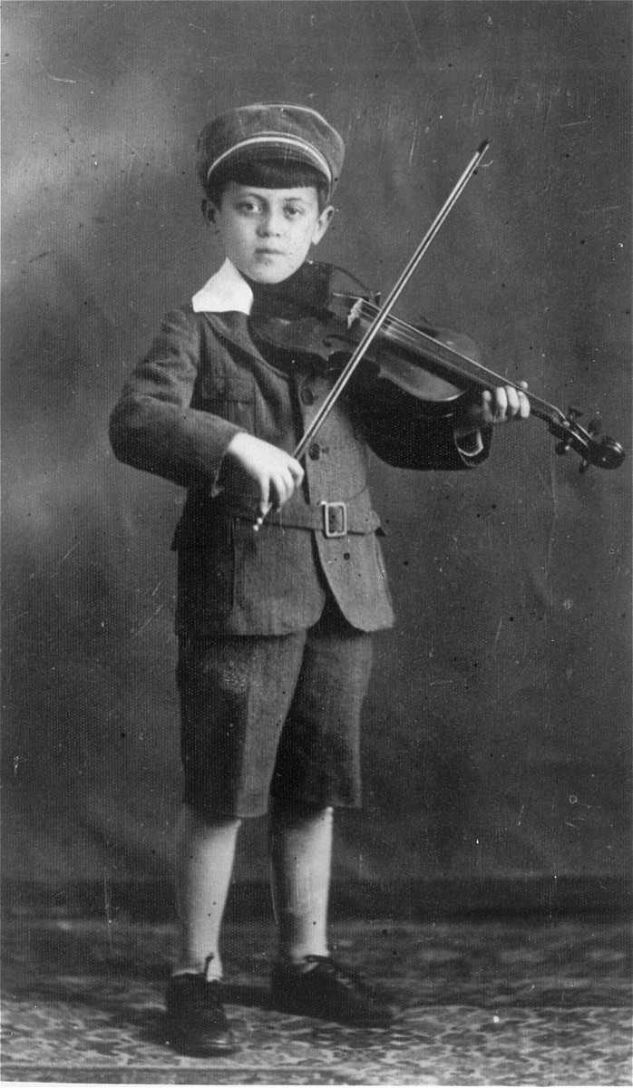 Portrait of a small child with dark shirt hair and light skin tone playing a violin. He stands looking at the camera while wearing a jacket, shorts, and hat.