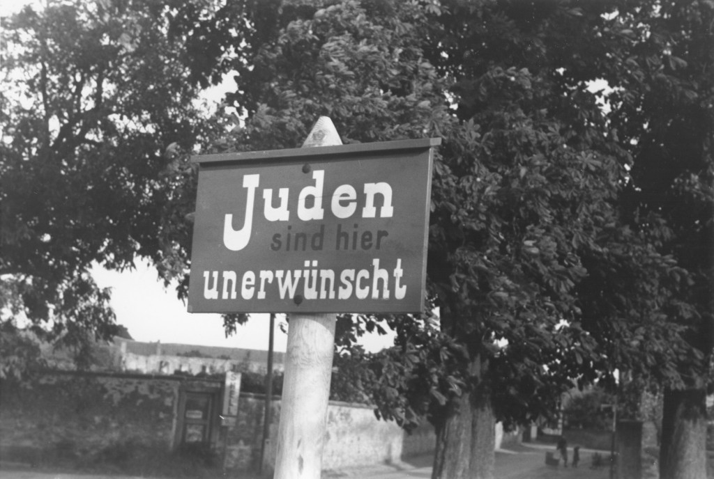 Anti-Jewish sign on a wooden post. Trees and homes can be seen in the background.