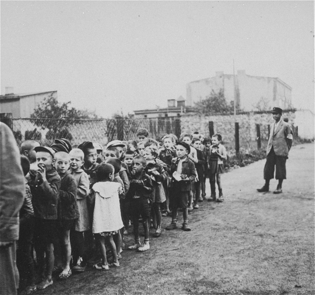 A large group of children gather in a dirt street. One adult with dark skin tone stands to one side looking at the group. The arm of another adult can be seen on the left side of the photograph.