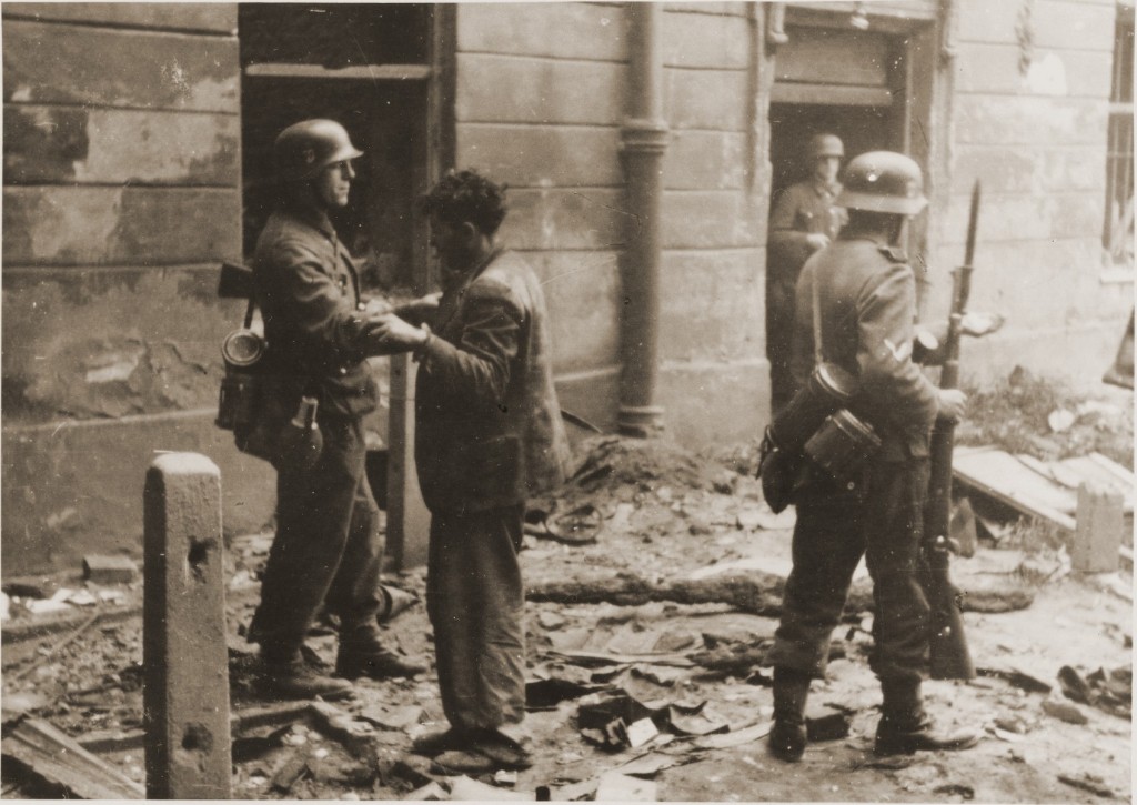 A disheveled man stands on a city street in front of a uniformed soldier surrounded by rubble. The soldier has his hand on the man’s shoulder and appears to be leading him. There are two other uniformed soldiers on the right side of the photograph. Both hold firearms.