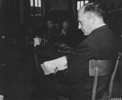Friedrich Hoffman, holding a stack of death records, testifies about the murder of 324 Catholic priests who were exposed to malaria during Nazi medical experiments at Dachau concentration camp.
