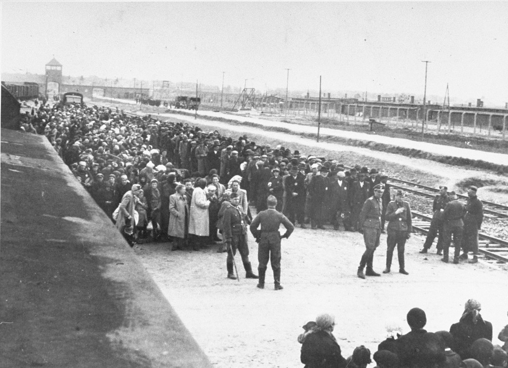 A large group of people in two groups stand near empty train tracks. Guards survey the groups. The Auschwitz II-Birkenau gatehouse can be seen in the background.