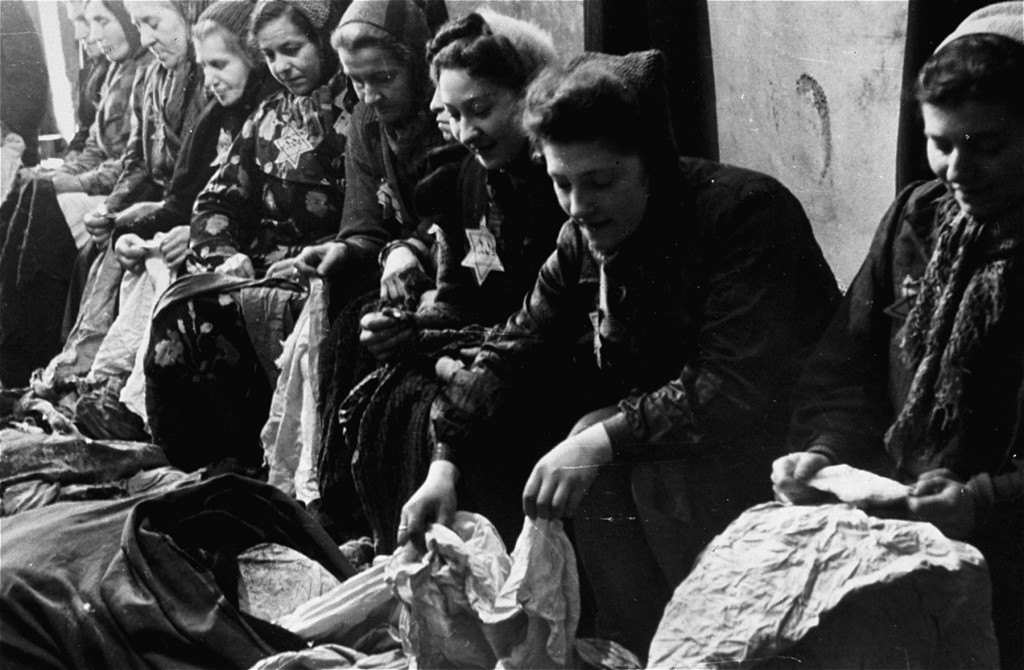 Ten women sit on a bench sorting through large sacks of clothing. Most of the women are wearing striped uniforms and head coverings. A few are wearing a Star of David patch on their chests.