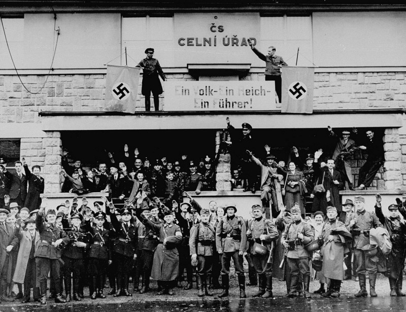 German troops marching into the Sudetenland stop at a former Czech frontier post. Nazi officials and Sudeten Germans salute the troops. The sign between the swastikas reads: "One People, One Reich, One Führer." Grottau, Czechoslovakia, October 2 or 3, 1938.