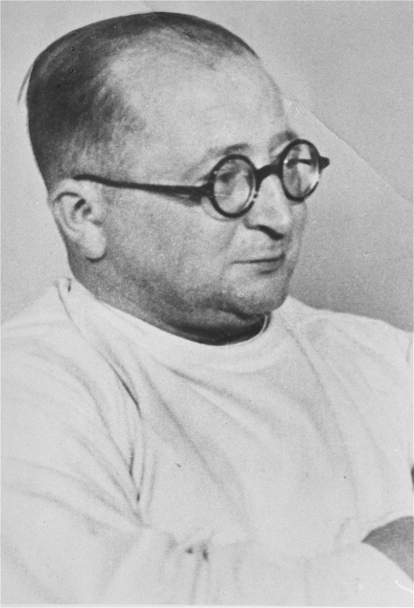 Portrait of a balding heavyset man with light skin tone. He is turned at an angle and appears to have his arms crossed. He wears a dark pair of round glasses and a white doctor’s uniform.