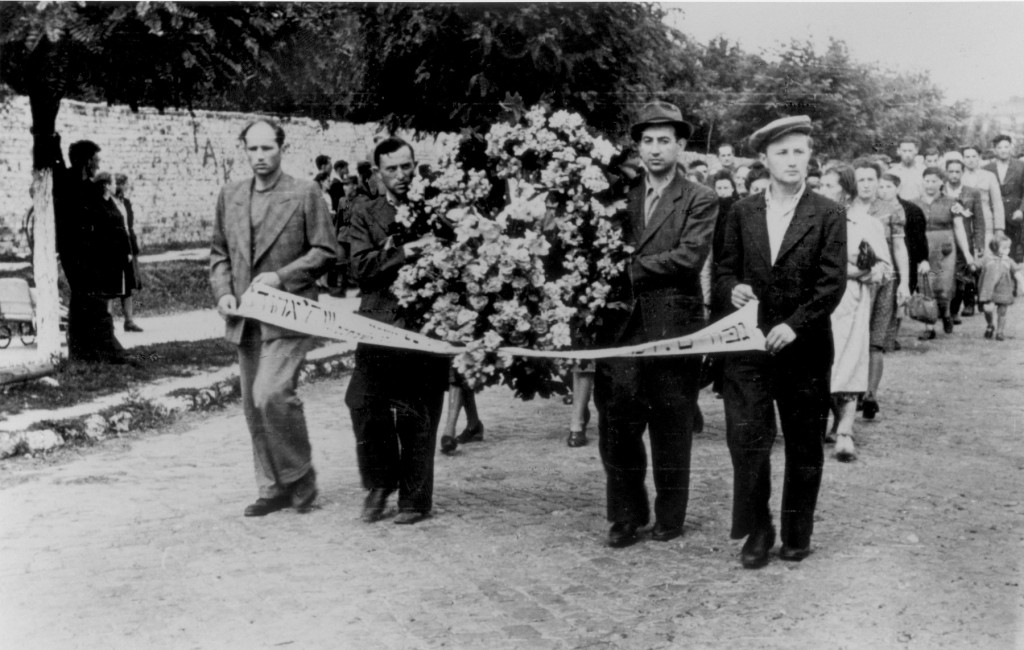A large group of people walk down a street in a funeral procession. Four men stand at the front carrying a large arrangement of flowers and a small banner.