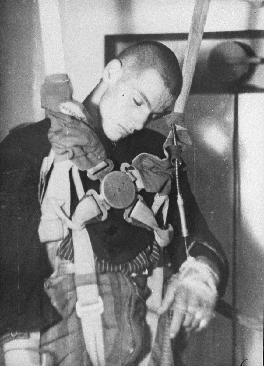 A male prisoner sits with a harness with testing equipment wrapped around him. The prisoner wears a uniform, with dark hair shaved close and light skin tone. He appears to be unconscious, with his head angled to the side and limp arms.