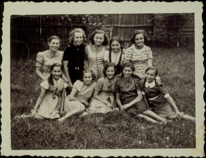 A black and white photograph with scalloped edges. Ten young girls with lighter skin tones form two rows, some sitting in the grass, some kneeling. All the girls are smiling at the camera.