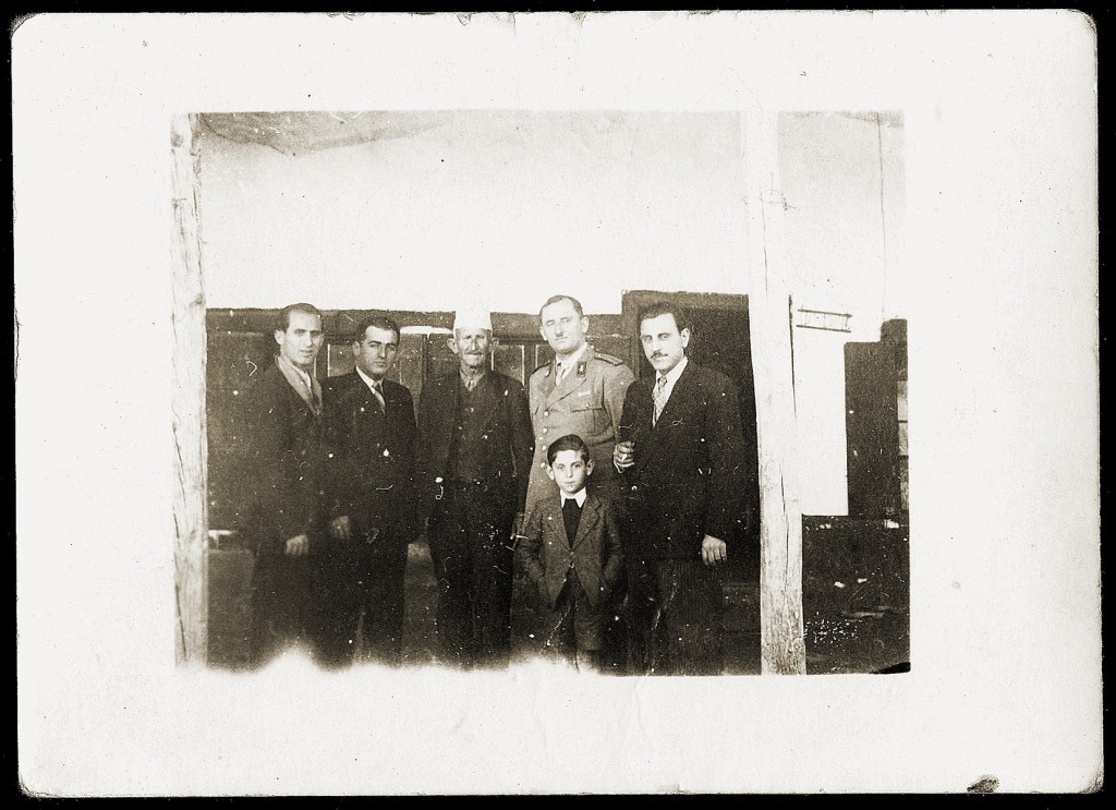 <p><span style="font-weight: 400;">Mosa (right) and Gavra (front) Mandil with local officials in Albania, including the mayor and chief of police of Kavaja, 1942–1943. </span></p>
<p><span style="font-weight: 400;">Jewish refugees were required to check in daily at the police station in Kavaja but were otherwise permitted to live freely in Albania. They felt welcomed by Albanian citizens. Albanian cultural traditions and ethical codes known as <em>Besa</em> </span><span style="font-weight: 400;">taught that a person must help somebody in need regardless of their faith or national origin. Albanian villagers brought the Jewish refugees food and blankets. Gavra Mandil would later recall that the warm welcome they received made his family feel “as if we have come to paradise.” </span></p>
<p><span style="font-weight: 400;">The Mandil family rented an apartment in Kavaja with several other Jewish refugee families. Mosa hung a sign from the upstairs balcony advertising his photography services. Gavra, who was nearly 7 years old, began attending school. Albania’s population was mostly Muslim, although Gavra had both Muslim and Christian classmates. He enrolled under a false identity, pretending to be a Muslim boy named Ibrahim Mele. Gavra learned Muslim prayers so people would not suspect he was Jewish. </span></p>