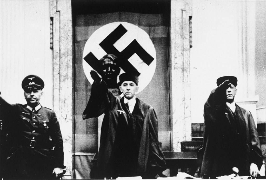 Three men stand with arms raised in Nazi salutes. There is a bust of Hitler on a pedestal behind them and a Nazi flag is hung on the wall.
